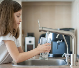Fototapeta na wymiar Portrait of a little caucasian girl gaining a glass of tap clean water. Kitchen faucet. Cute curly kid pouring fresh water from filter tap. Indoors. Healthy life concept
