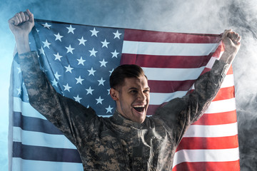 happy soldier gesturing while holding american flag on black with smoke