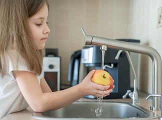 A cute little girl washes a red yellow apple in a sink. Child is washing the fruit in kitchen. Little child learns the rules of hygiene. Child uses the tap himself. Healthy lifestyle hygiene concept