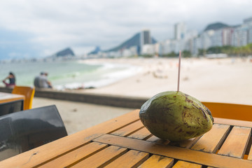 Whole coconut with straw on a wooden table with a view of Copacabana beach in a bright day light, Rio de Janeiro, Brazil.