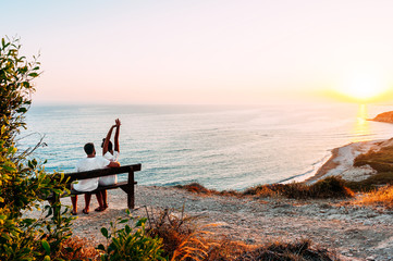 Happy couple at sunset. Man and woman at dawn. Couple sitting on a bench by the sea. Couple in love kissing by the sea. Man and woman traveling. Honeymoon trip. Sunset on sea. Dawn by the sea