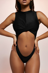 Obraz premium Cropped front view shot of African lady, wearing ink black one-piece silky swimsuit with front cut-out. The girl is posing with her hands on waist, sliding down to hips on light purple background.