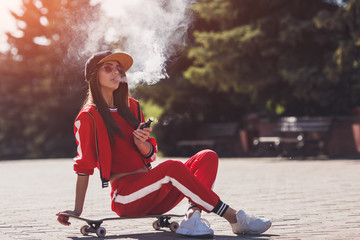Vaping girl. Young woman sitting on skateboard and vape e-cig. Pretty young female in black hat,...