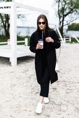 The young pretty brunette girl spends time alone on the beach at the lake. Spring. The teenage girl holds a glass of paper with her drink in her hands