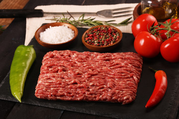 raw minced meat on plate, ingredients for burger with tomato, pepper, onion, spices and seasonings on black background.