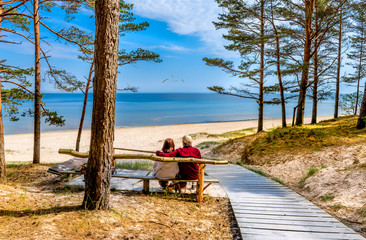 Happy couple of seniors are resting and looking at a distance near a sandy beach of the Baltic Sea - obrazy, fototapety, plakaty