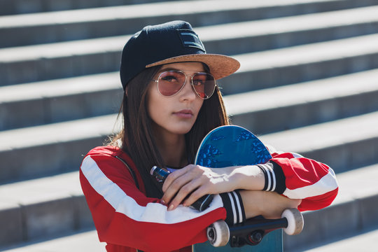 Vaping girl. Young woman with skateboard vape e-cig. Pretty young female in black hat, red clothing vape ecig, vaping device at the sunset. Toned image. Hip-hop style.