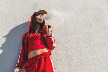 Vaping girl. Young woman standing in front of a white wall and vape e-cig. Pretty young female in...