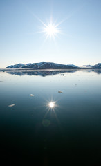 A sunburst in the sky is reflected in still arctic waters with arctic land on the horizon