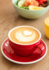 A red cup of cappuccino with trendy latte art on a saucer with a coffee spoon. In the background a healthy breakfast bowl. 