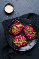 Bruschetta with beetroot hummus, arugula and sesame seeds on dark concrete background. Top view.