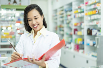 Cheerful Asian woman pharmacist checking medicines inventory at hospital pharmacy. Pharmacist in drugstore or pharmacy taking notes. Portrait of health care doctor in pharmacy writing on clipboard