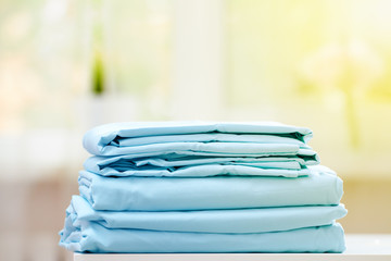 Close-up of blue clean bedding on a blurred background. A stack of folded new bed sheets on the table. Sunlight from the window.