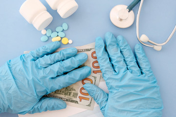 Hands in blue gloves on an envelope with American hundred-dollar bills. Top view of the doctor's table, stethoscope, pills. The concept of receiving payment for services, paid medicine, corruption.