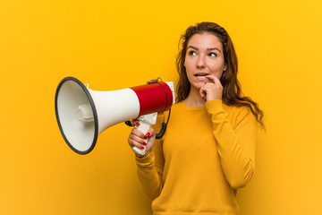 Young european woman holding a megaphone relaxed thinking about something looking at a copy space.