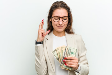 Young european business woman holding dollar banknotes crossing fingers for having luck
