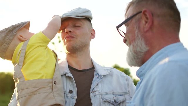 Male three generations of the same family for a walk. One year old child plays with his father's cap. Sun glare