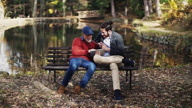 Senior father and his son sitting on bench in nature, using tablet.