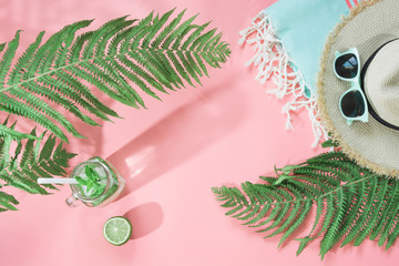Straw beach hat, sunglasses, towel, leaf of fern and refreshing drinks with lime on pastel pink background. Top view. Summer tropical vacation.