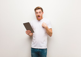 Young redhead man surprised, feels successful and prosperous. Holding a tablet.