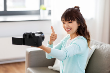 Fototapeta na wymiar blogging, videoblog and technology concept - asian woman or blogger with camera recording video blog or taking selfie and showing thumbs up at home