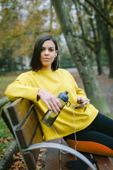 Sporty young woman on outdoor autumn fitness workout taking a break for drinking water and listening music from smart phone. Female athlete sitting on a park bench using cell phone.