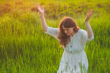 Naklejka na ściany i meble Girl closed her eyes, praying in a field during beautiful sunset. Hands folded in prayer concept for faith, spirituality and religion. Peace, hope, dreams concept