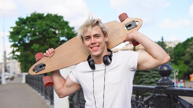 Cheerful blonde man with headphones holding longboard behind head and smiling