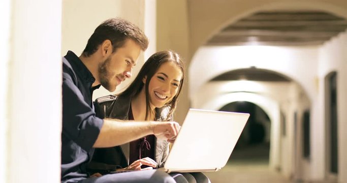 Happy couple browsing laptop content sitting in a town street in the night