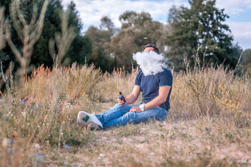 Naklejka na ściany i meble Modern man is resting in nature and vaping and letting off steam from an electronic cigarette. Vaping activity.