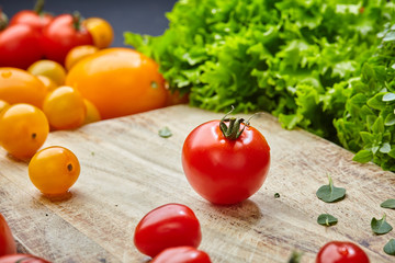 Ripe cherry tomato on a wooden cutting board surrounded by lettuce, basil leaves and yellow tomatoes. Concept of cooking healthy food. Colorful still life