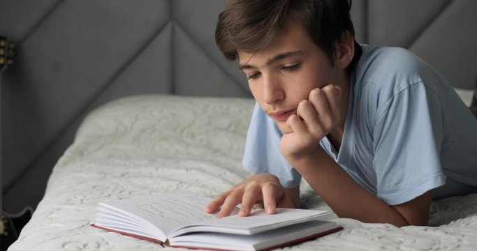 Young man reading book at home  lying on bed. Teenage child reads book in lunch time. Teen boy guy with a book lies on a sofa at afternoon . Real time. 4k footage.