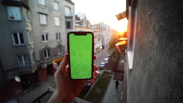 Close-up of man looking at smartphone sitting on the balcony with beauty sunset above the street. First-person view of male hand with iPhone.