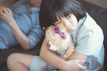 Asian female kid sit on couch hug a bear sad looking outside while her father playing phone don't care for his daughter. Asian problem child girl depress from seeking attention from parents