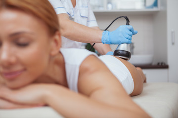 Selective focus on ultrasound cavitation machine professional cosmetologist is using on female client. Woman receiving anti-cellulite slimming procedure at beauty salon - obrazy, fototapety, plakaty