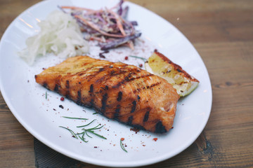 Delicious grilled salmon fish steak served with seasonings, veggies and lemon on a white plate on a wooden table. Restaurant menu design. Barbecue party. 