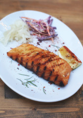 Delicious grilled salmon fish steak served with seasonings, veggies and lemon on a white plate on a wooden table. Restaurant menu design. Barbecue party. 