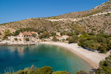 Dubovica beach in the Adriatic Sea on the island of Hvar. The pebble beach is sheltered in an emerald green lagoon set against a small cluster of historic stone houses nestled among rolling hills.