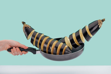 Cropped view of woman holding frying pan with freshly sliced organic eggplants above white table isolated on turquoise