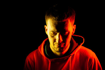 An adult guy stands with his head tilted down in an orange hooded sweatshirt highlighted in red and yellow on the sides with a pensive and regretful view on a black isolated background.