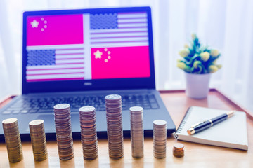 USA vs China flag in laptop screen on wooden table. Gold and silver coins are front of laptop and notebook with pen.