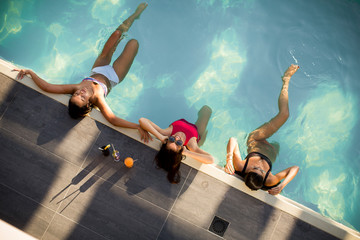 Three beautiful slim women in bikini relaxing and drink cocktails on poolside of a swimming pool - obrazy, fototapety, plakaty