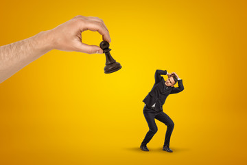 Little scared businessman covering his head with hands to protect from huge hand holding black chess pawn on yellow background.