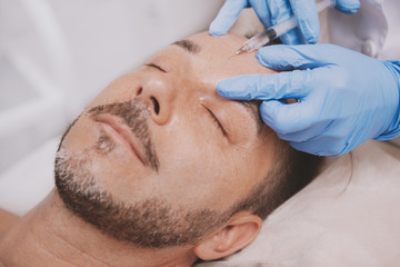 Obraz na płótnie Canvas Close up of a handsome mature bearded man receiving face injections at beauty clinic. Professional cosmetologist doing wrinkle filler injections for male client