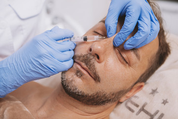 Mature man getting liquid rhinoplasty by professional cosmetologist. Attractive male client getting non-surgical nose job at cosmetology clinic. Beautician injecting filler in the face of mature man