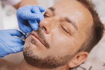 Beaeded mature handsome man getting anti-wrinkle treatment at beauty clinic. Professional cosmetologist doing facial filler injections for male client