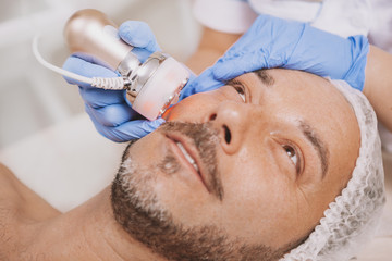 Happy handsome mature man receiving facial hardware cosmetology treatment at beauty salon. Relaxed attractive man getting microdermabrasion procedure by professional cosmetologist