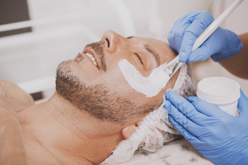 Mature bearded joyful man receiving facial spa treatment, beautician applying mask on the face of...