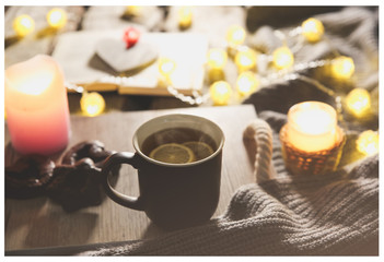 Holidays mood photo. Christmas lights and warm knitted sweater. Hot tea mug with a book for cosy evening. Sweet gingerbread and wooden heart on tray. Perfect winter flat lay with candle. Hygge concept