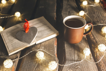 Holidays mood photo. Christmas lights and hot tea mug. Book for cosy evening. Sweet gingerbread and wooden heart on tray. Perfect winter flat lay. Hygge concept.